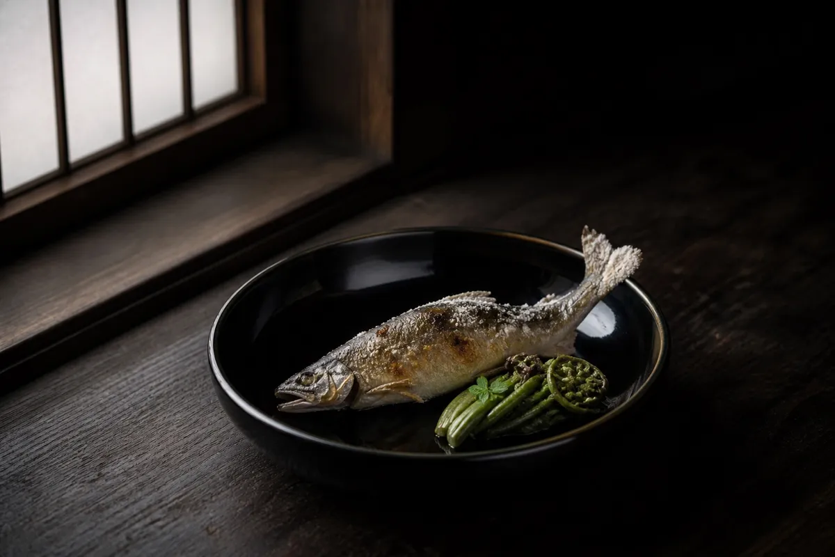 黒漆の器に盛られた川魚の塩焼きと旬の山菜。窓からの自然光がひと筋差す（イメージ）